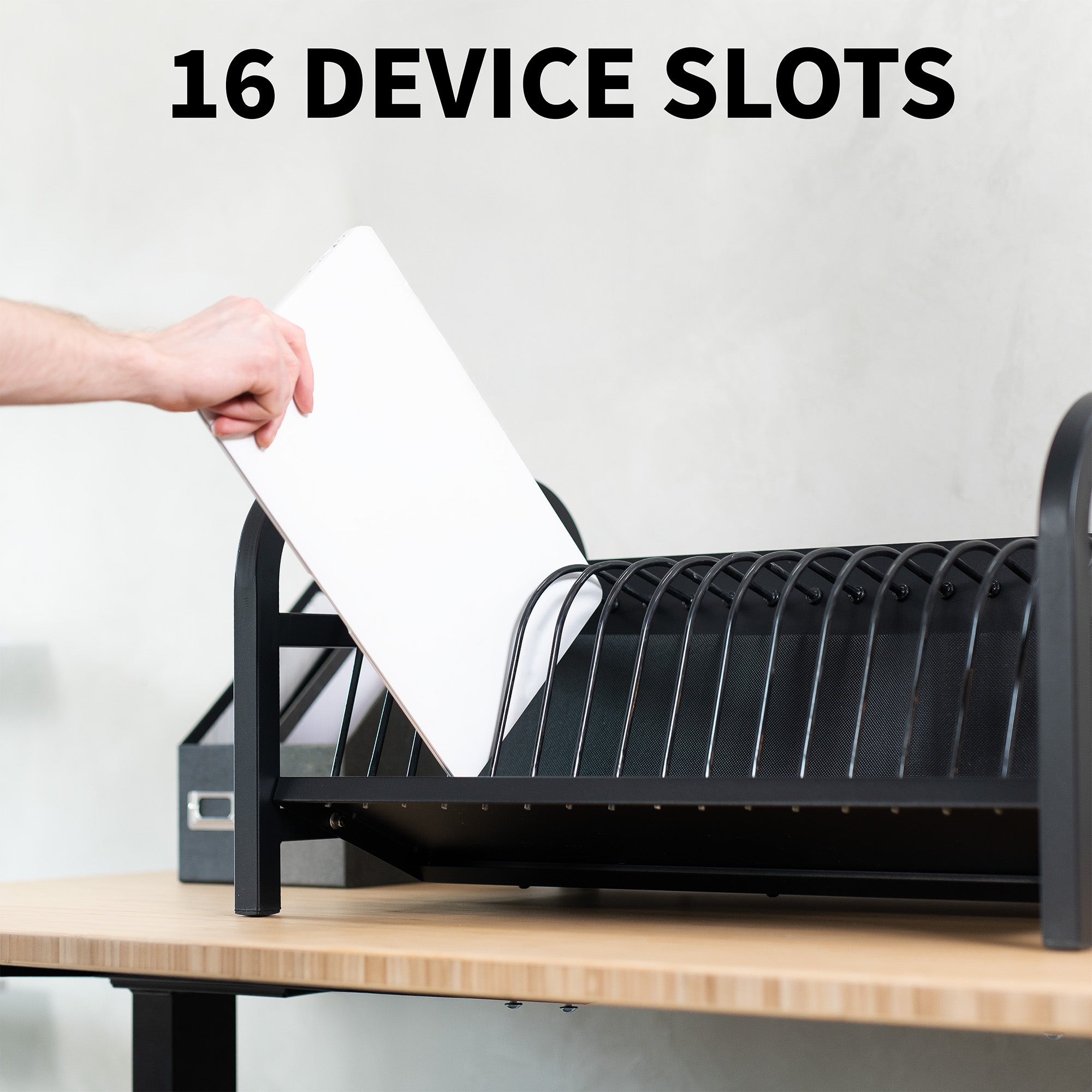 Black charging organizer with 16 device slots on a wooden surface, with a hand placing a laptop into it.