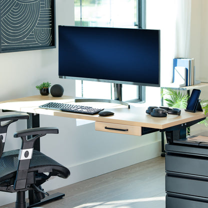Modern office desk setup with a computer monitor, keyboard, and light wood storage drawer in a bright room.