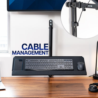 Desk setup with keyboard on wall-mounted keyboard tray and a wall-mounted monitor on a wooden desk with cable management solution.
